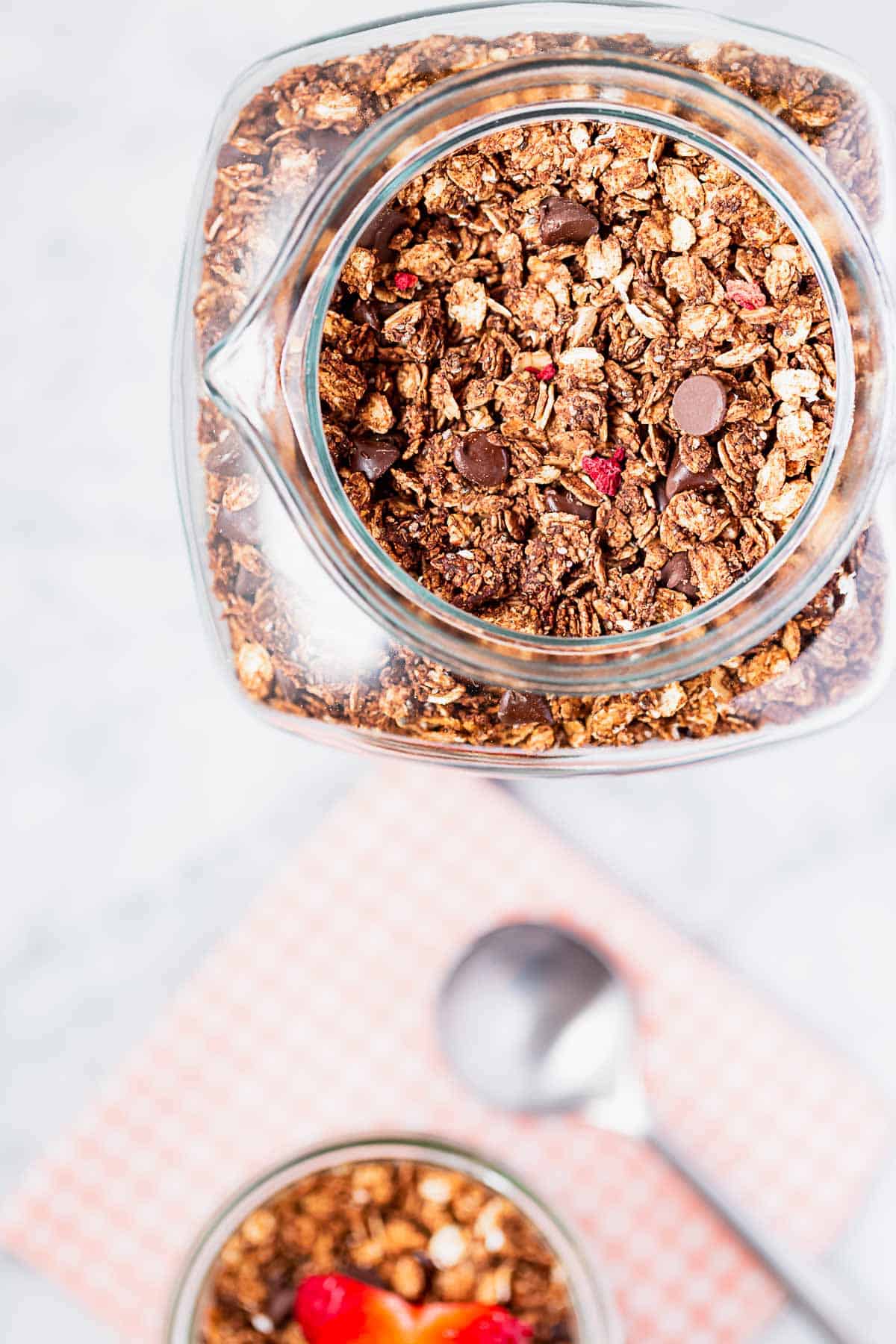 A large jar of homemade chocolate berry granola next to a parfait.