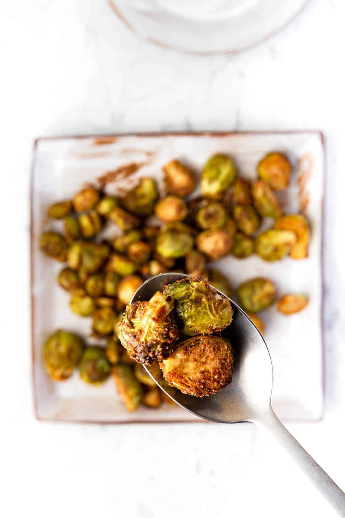 A square plate of maple balsamic roasted brussels sprouts with a spoon holding some hovering above.