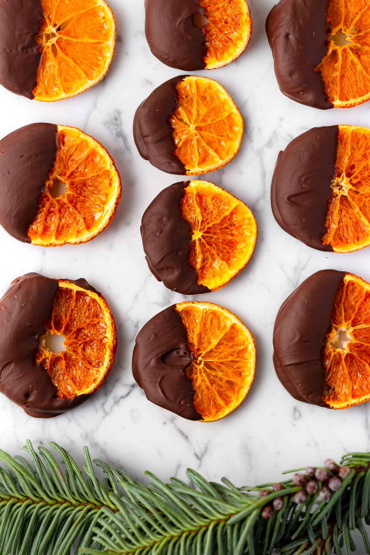 Chocolate-dipped satsuma orange slices on a marble surface with a Christmas tree branch below.
