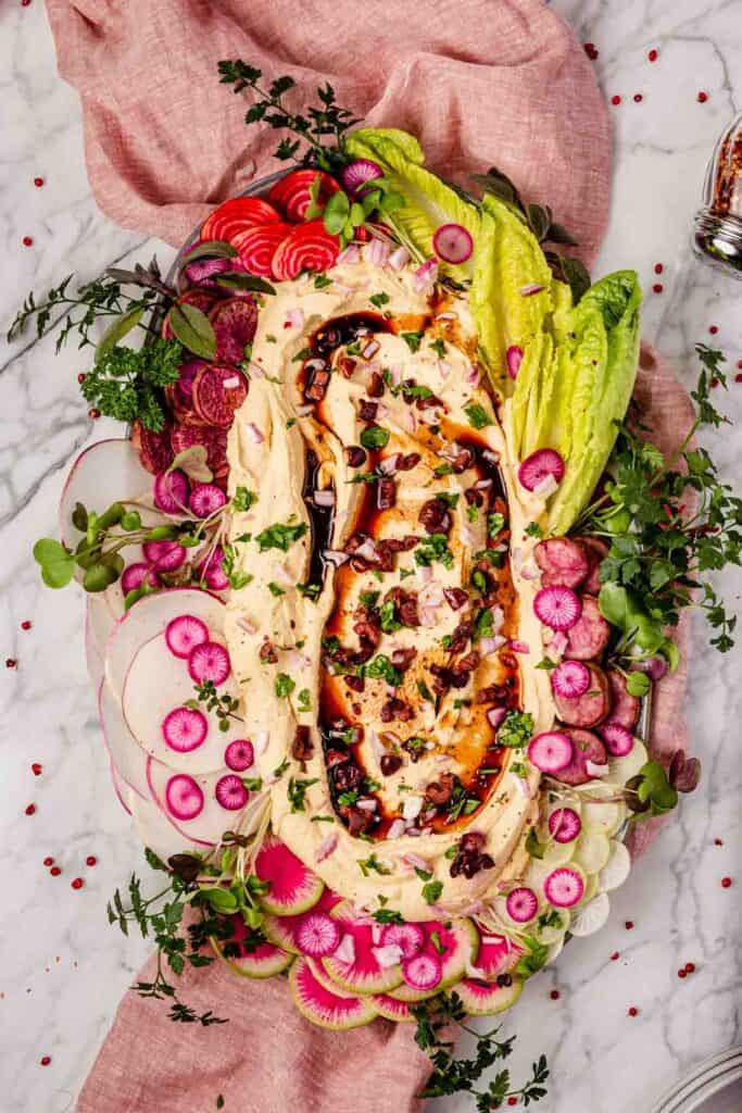 A crudite platter with creamy oil-free hummus at the center, topped with balsamic glaze and surrounded by colorful veggie slices