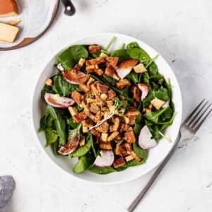 A bowl of spinach salad with tempeh bacon, red onion, and artisan vegan cheese