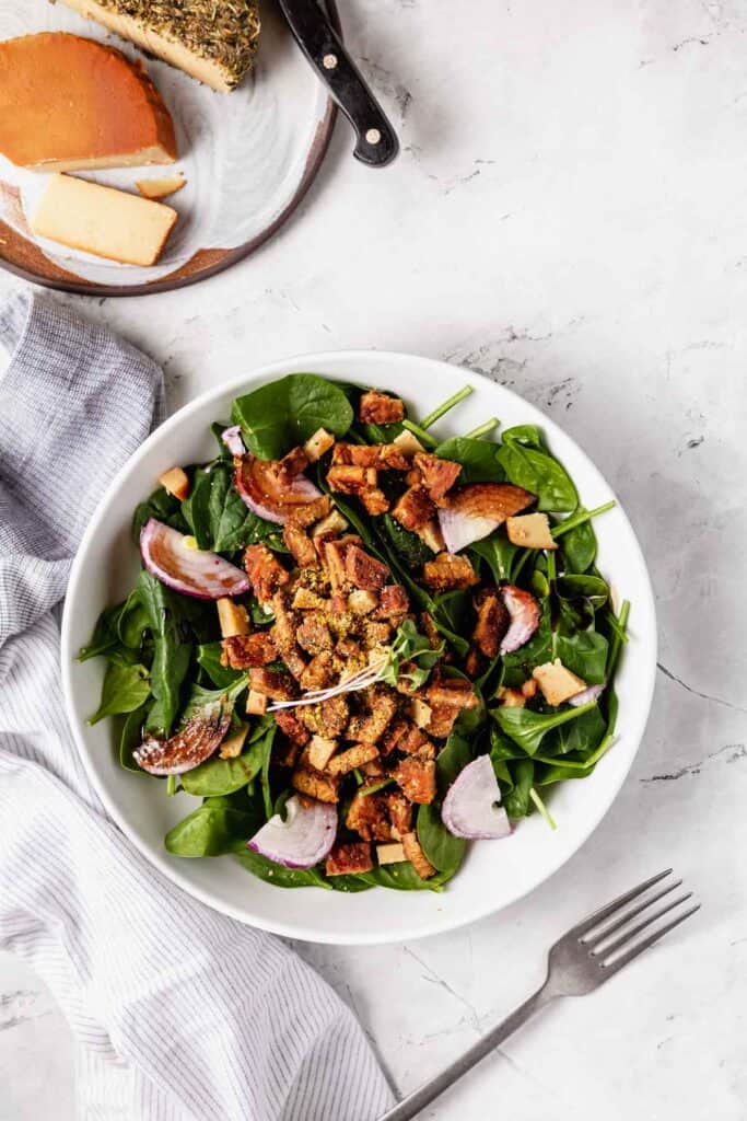 A big bowl of spinach salad topped with tempeh bacon pieces and vegan cheese