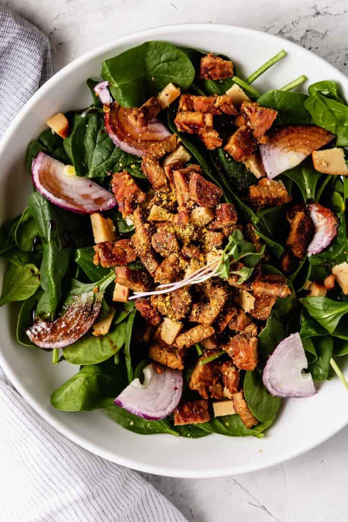 Close-up of a bowl of spinach salad with tempeh bacon and rainbow-shaped red onion