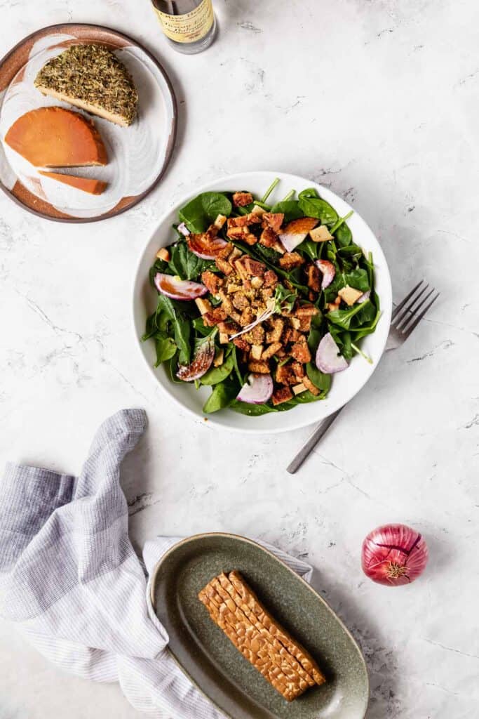 A plate of vegan artisan cheese above a bowl of spinach salad with tempeh bacon and an oblong bowl of tempeh bacon strips below