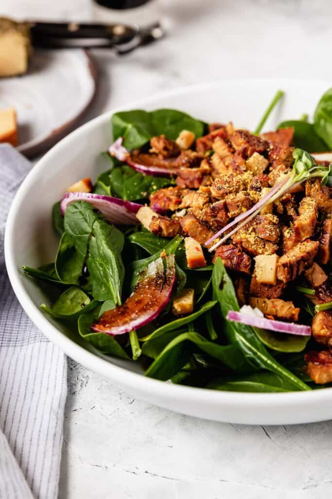 A side view of a big bowl of spinach salad topped with shaved red onion and tempeh bacon