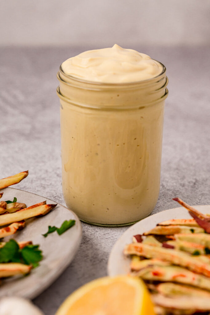 A tall jar of lemon garlic aioli surrounded by plates of fries.