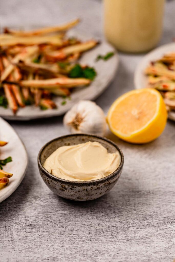 A small bowl of lemon garlic aioli with lemon and gqrlic in the background with fries.