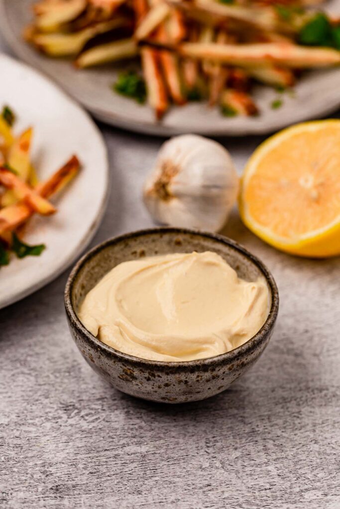 A bowl of vegan lemon garlic aioli surrounded by plates of fries, lemon, and garlic