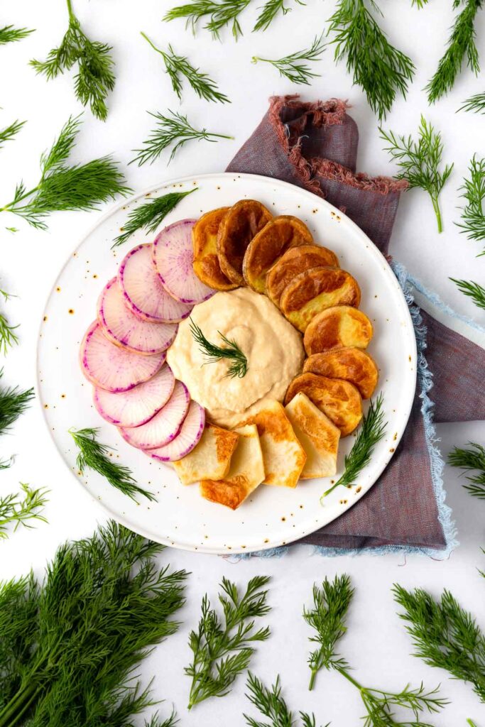 A plate of sliced purple radishes and potatoes surrounding hummus surrounded by fresh dill.
