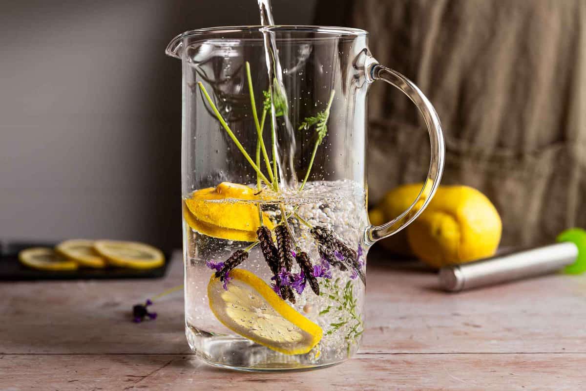 A water pitcher on a wood surface with lemon slices and lavender fronds being filled by a person with a linen apron on. 