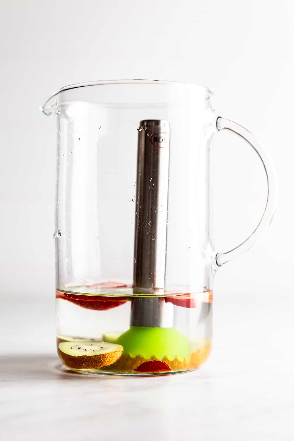 A glass pitcher with a muddler in it pushing on kiwi and strawberry pieces, with a little water in the pitcher.