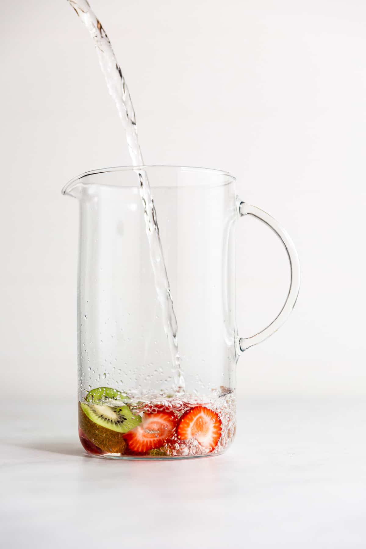 A glass pitcher with water pouring into it and sliced strawberries and kiwi at the bottom.