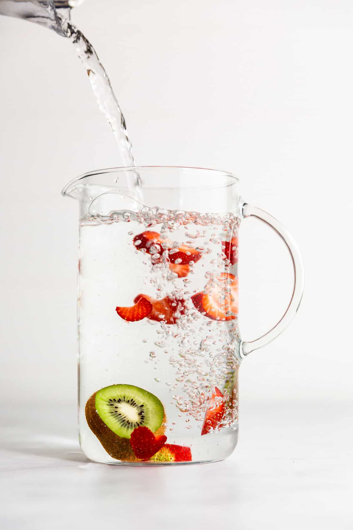 A pitcher of water with sliced strawberry and kiwi in it being filled.