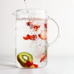 A glass pitcher filling with water, with kiwi and strawberry slices floating in it.
