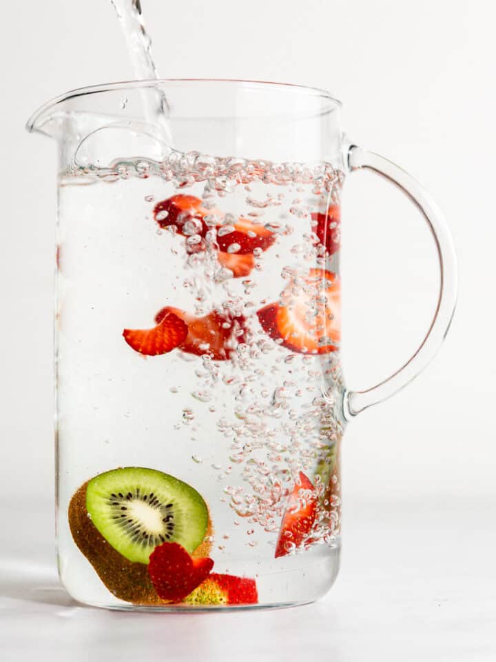 A glass pitcher filling with water, with kiwi and strawberry slices floating in it.