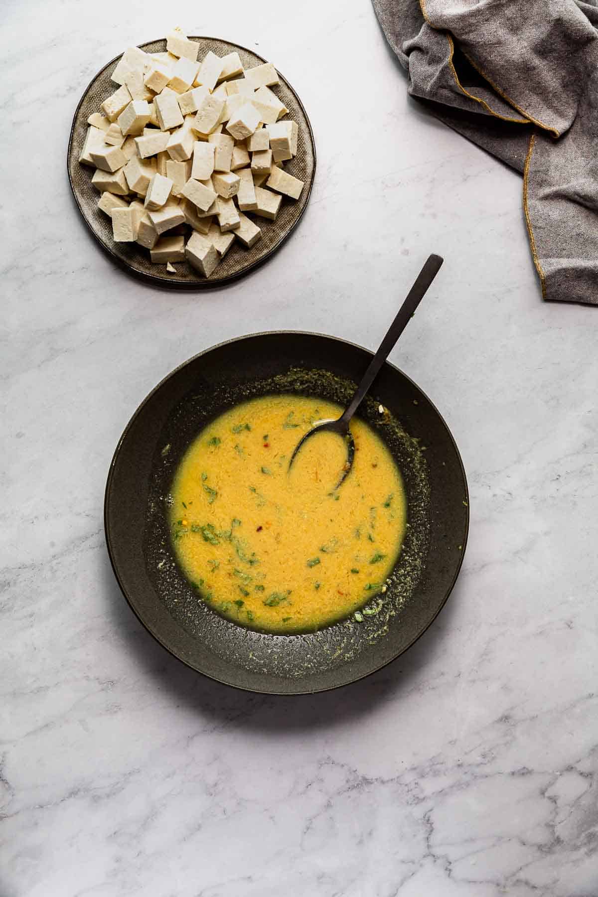 Chopped tofu on a plate above a dark bowl filled with golden marinade.