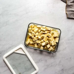 Tofu feta marinating in a glass container on a marble surface.
