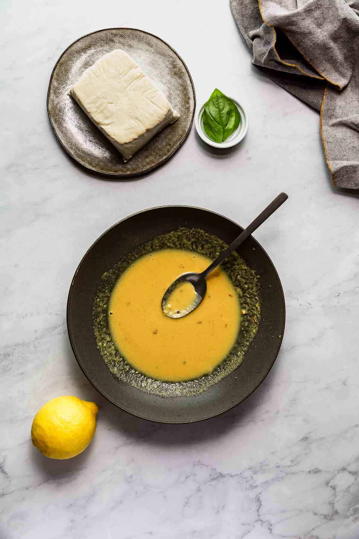 A block of tofu sitting above a dark grey bowl filled with golden marinade.