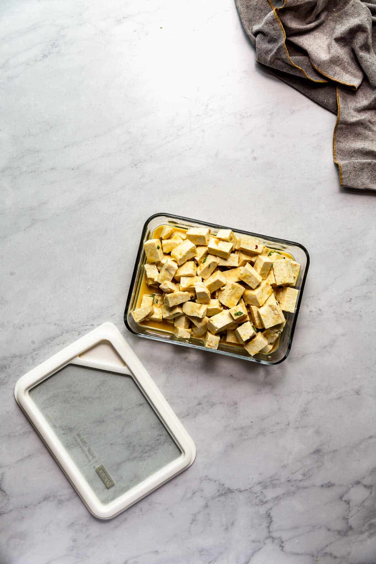 Bite-sized, marinated tofu pieces in a glass container on a marble surface.