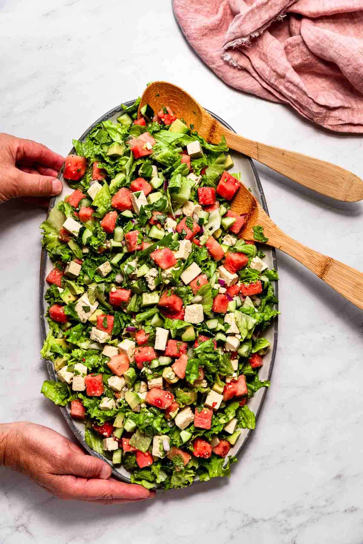 A man's hands placing a platter filled with watermelon salad onto a marble surface.