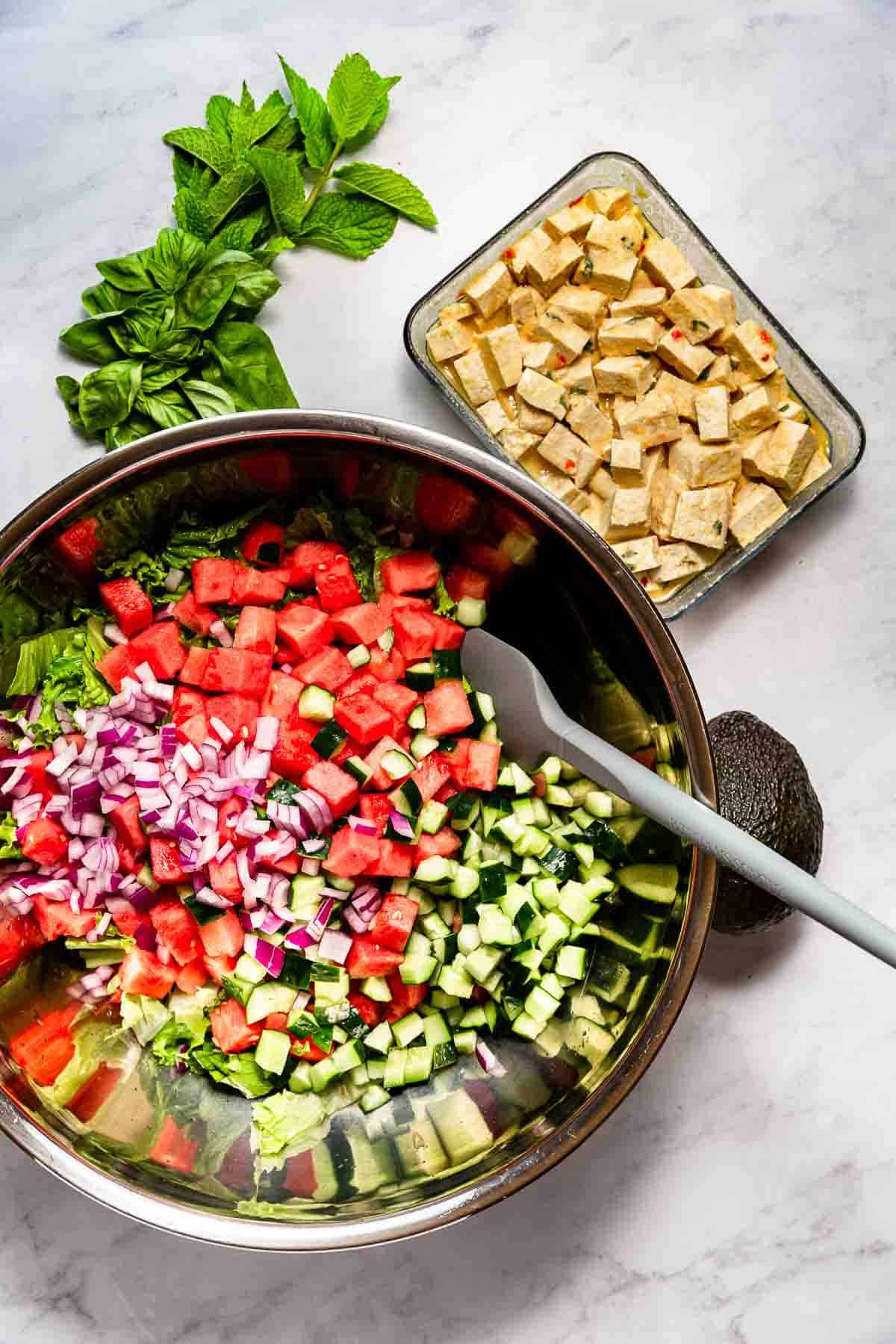 Salad ingredients for a vegan version of Kate Middleton's favorite in a large stainless bowl surrounded by fresh herbs and a container of tofu feta.