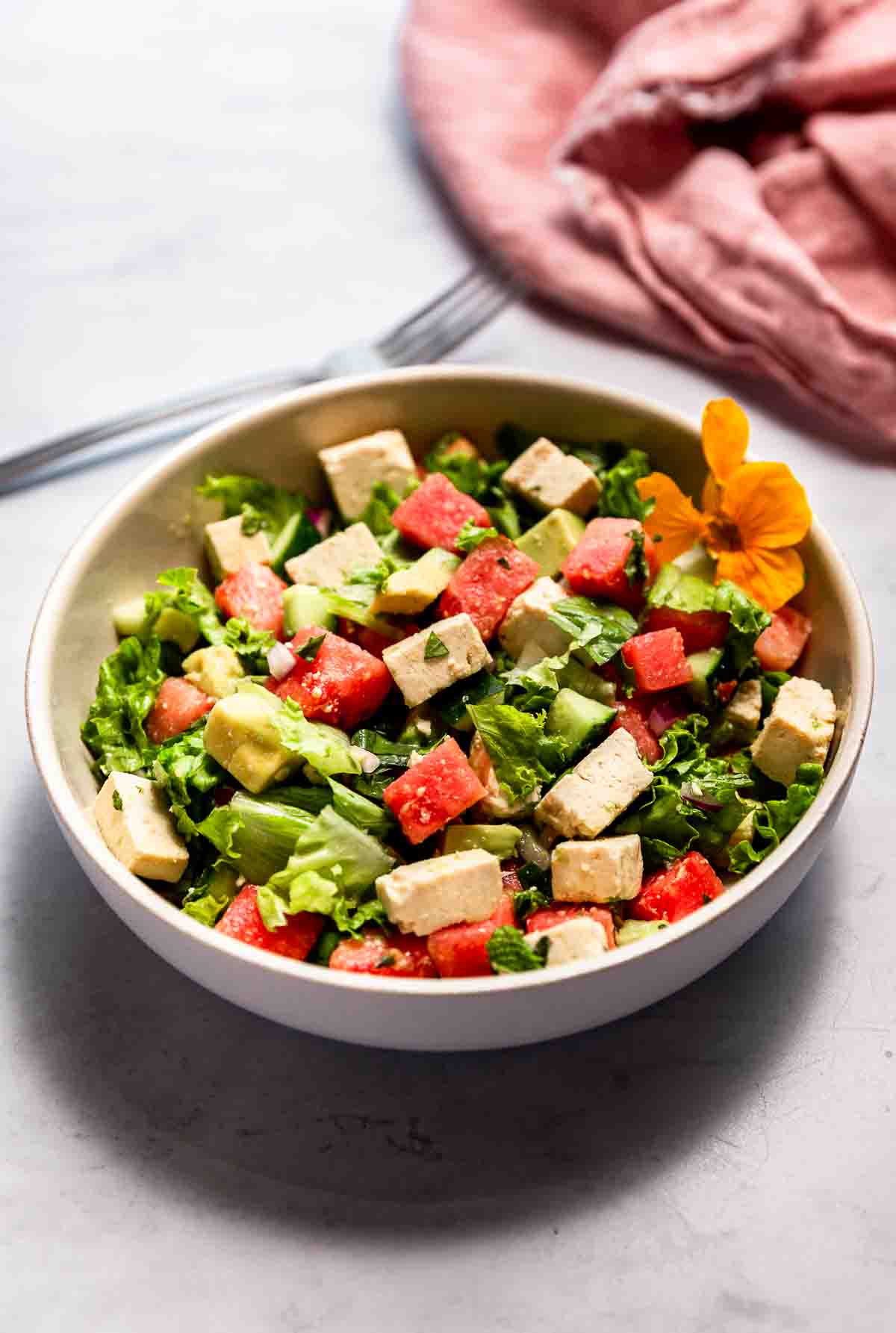 A closeup view of a bowl of salad with watermelon, tofu feta, cucumber, and herbs.