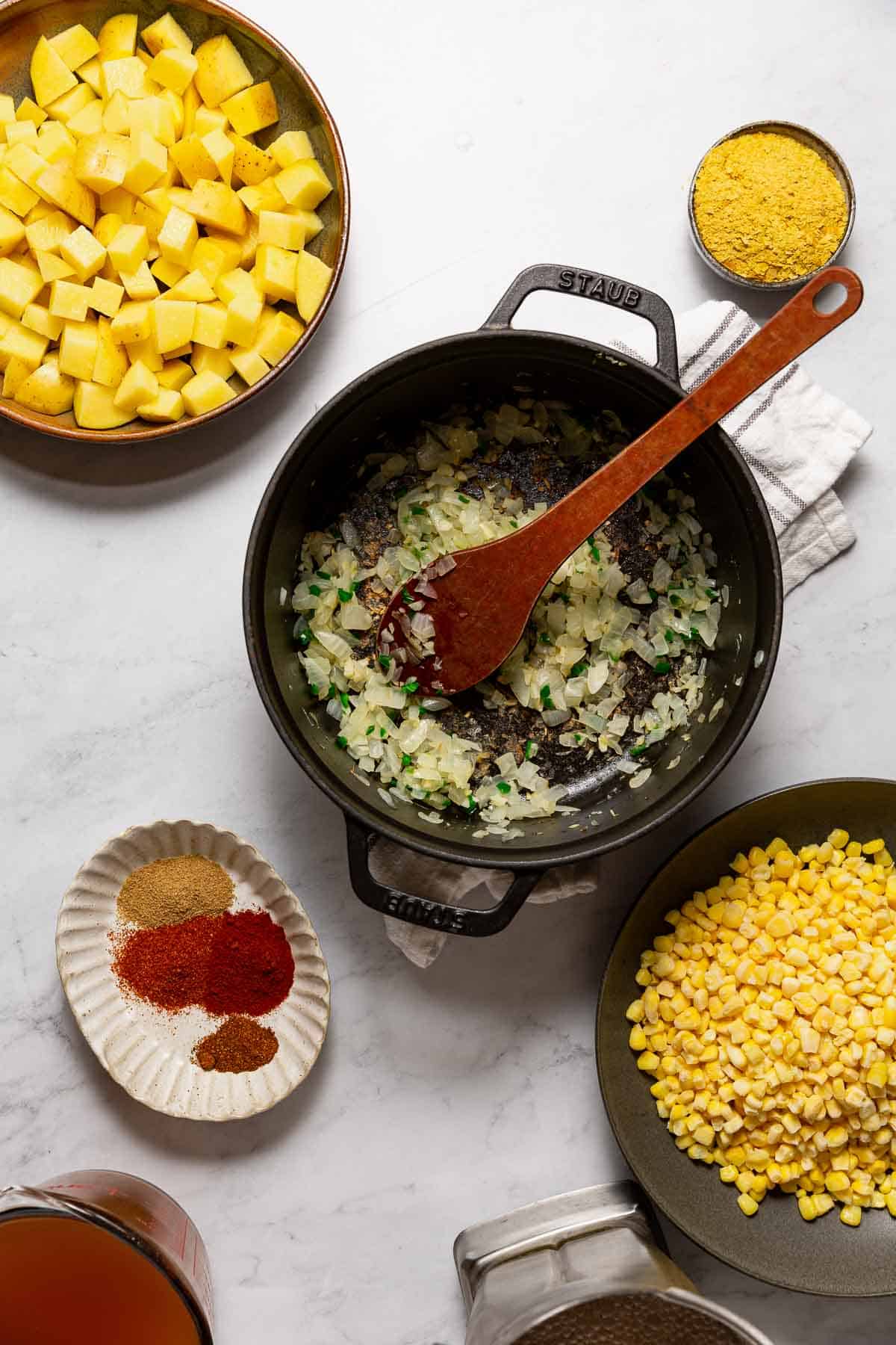 A large soup pot with translucent onion, garlic and jalapeno in it, surrounded by remaining ingredients for Easy Homemade Vegan Potato Corn Chowder.