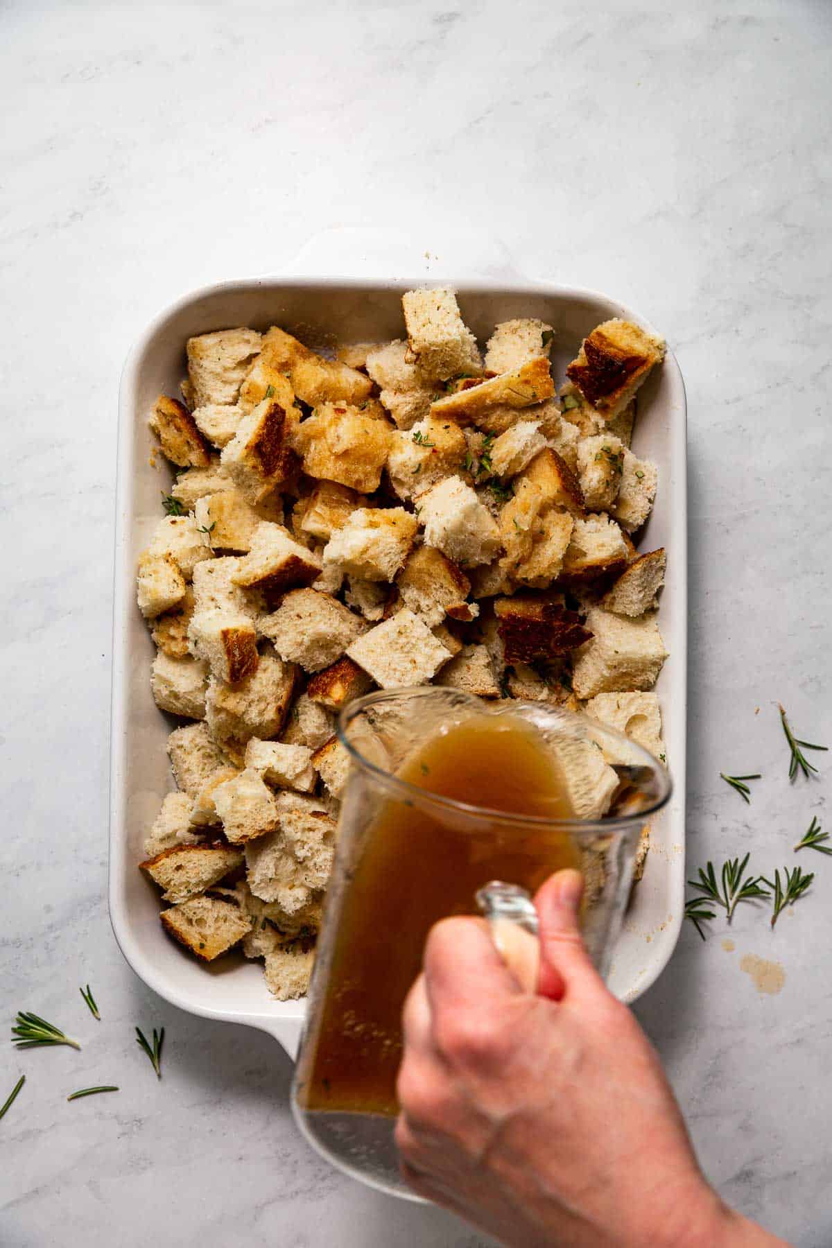 A hand pouring a pitcher of broth over a baking dish filled with large sourdough bread cubes and fresh herbs, making simple sourdough stuffing.