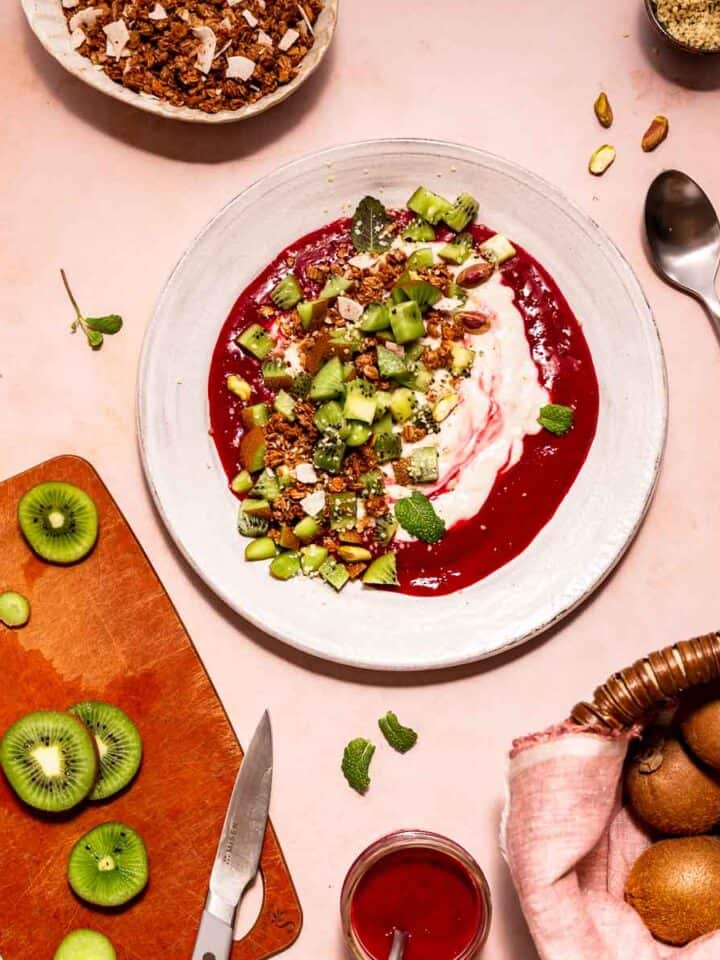 Chopped kiwi topping a plate of yogurt and cherry sauce with kiwi in a market basket and sliced on a chopping board surrounding the plate.