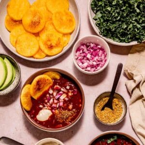 Toasted polenta rounds filling a large plate, surrounded by shredded kale, chopped red onion, and bowls of vegan chili.