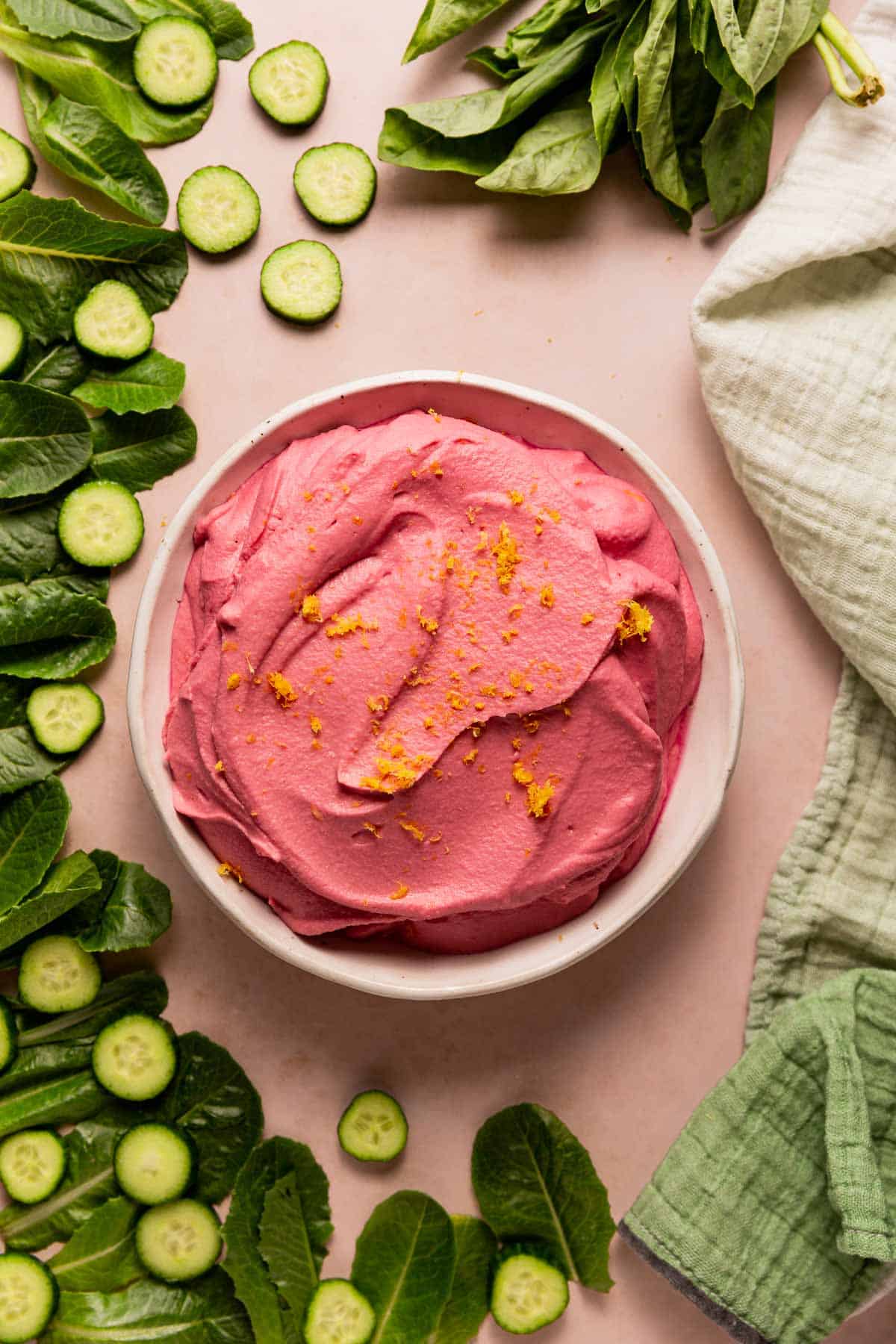 A big bowl of pink pickled beet hummus surrounded by lettuce, cucumbers, and basil.