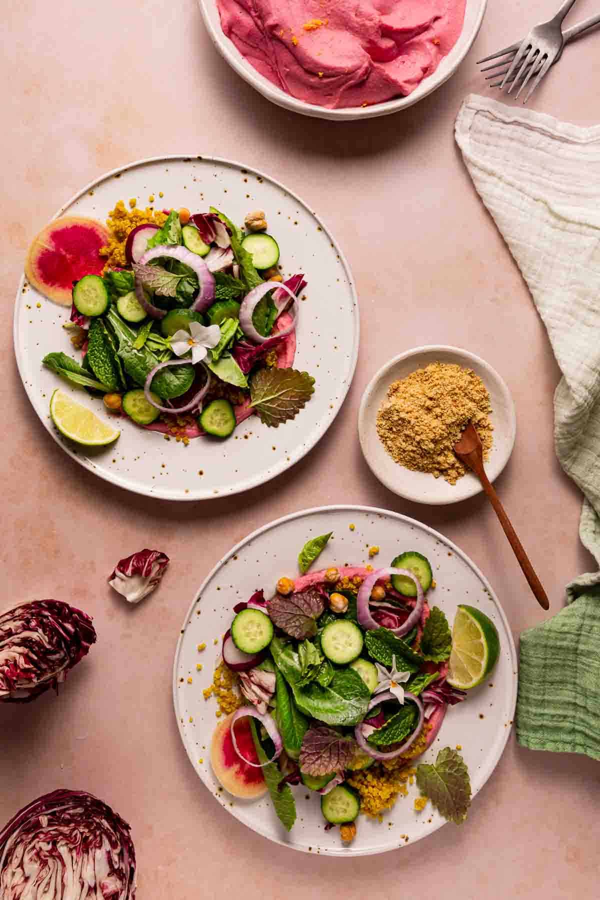 Two plates with pink beet hummus topped with colorful salads.