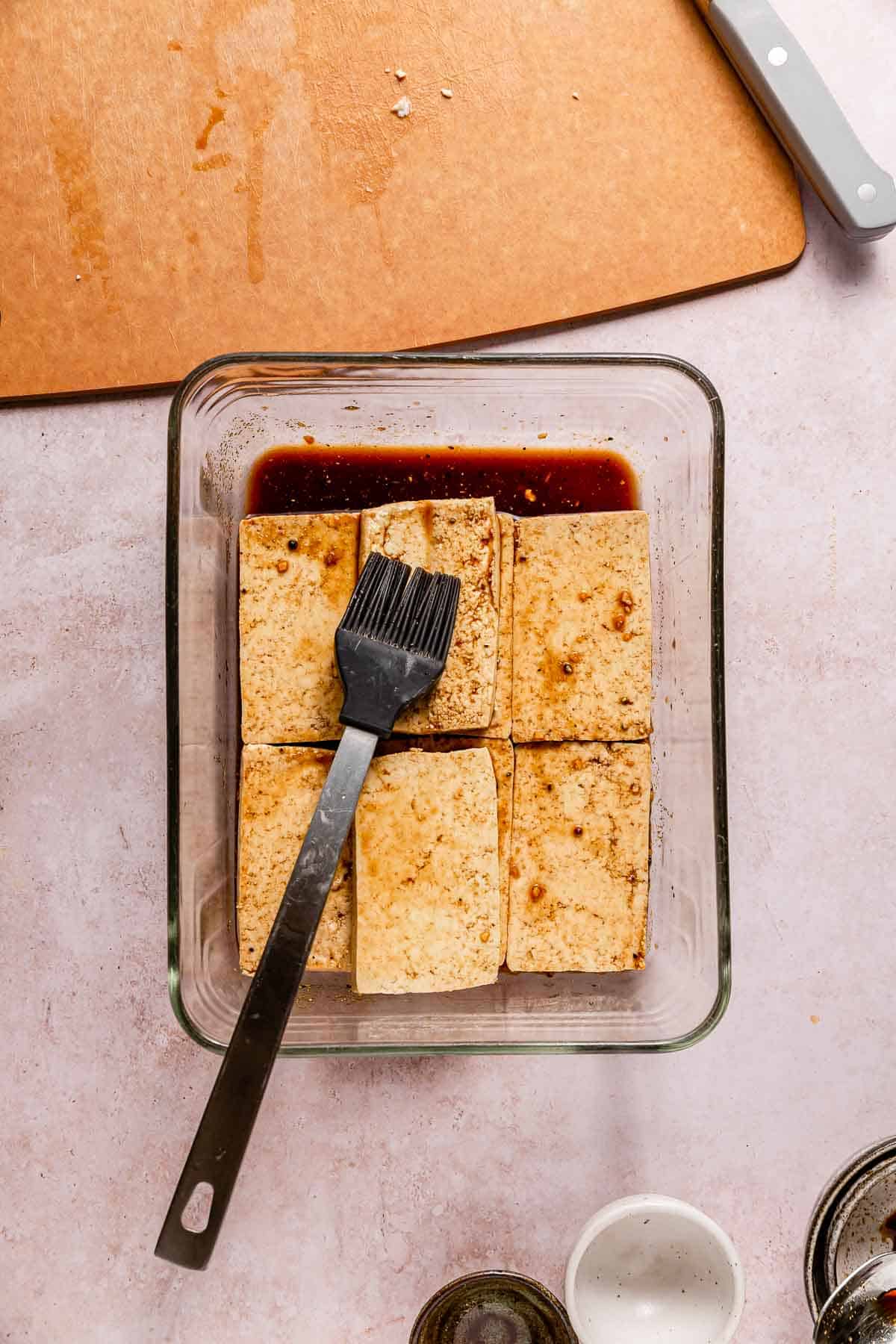 Tofu steaks in a container filled with marinade. A pastry brush is on top for brushing on the marinade.