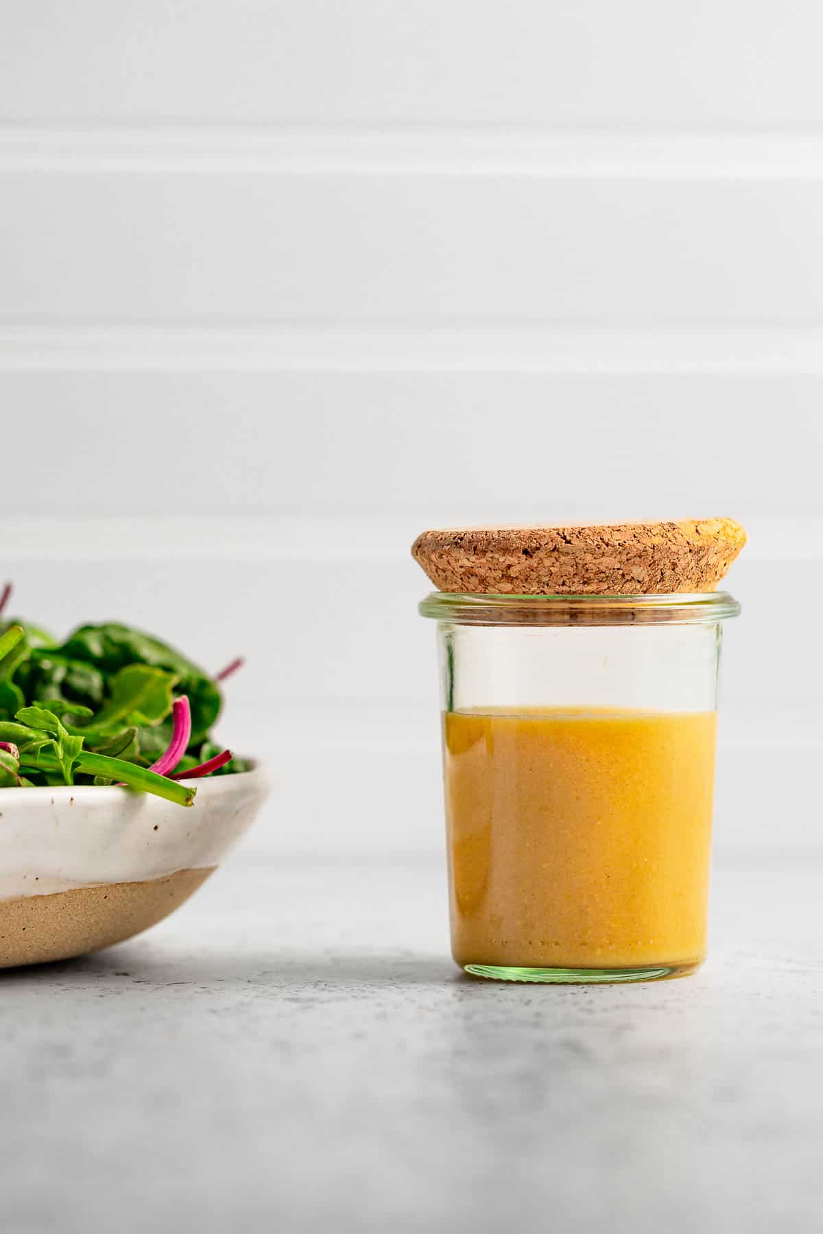 A jar of maple dijon dressing next to a green and pink salad.