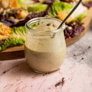 A jar of nut-free vegan salad dressing in front of a board containing caesar salad.