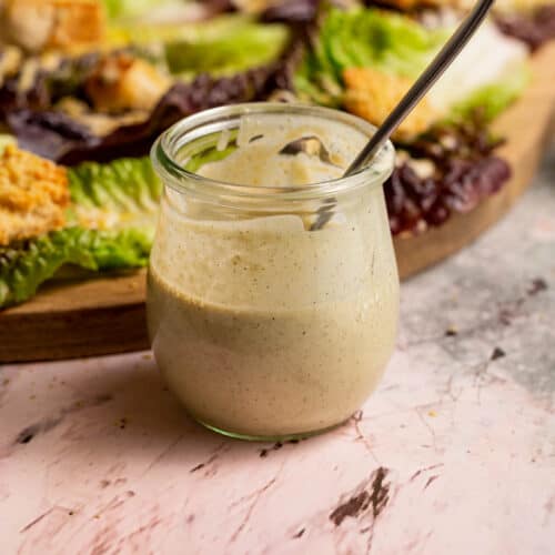 A jar of nut-free vegan salad dressing in front of a board containing caesar salad.