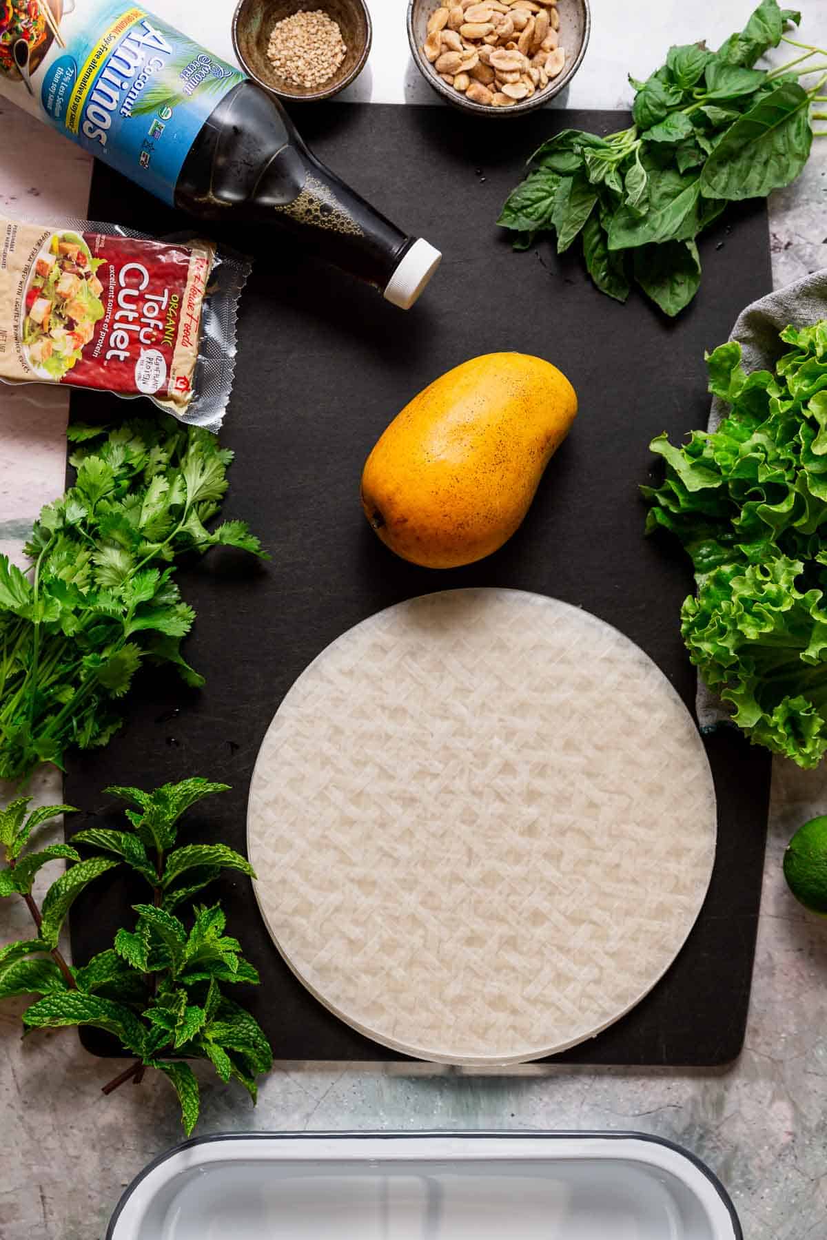 A mango on a black cutting board surrounded by rice paper wrappers, fresh herbs, tofu, nuts and seeds, and coconut aminos.