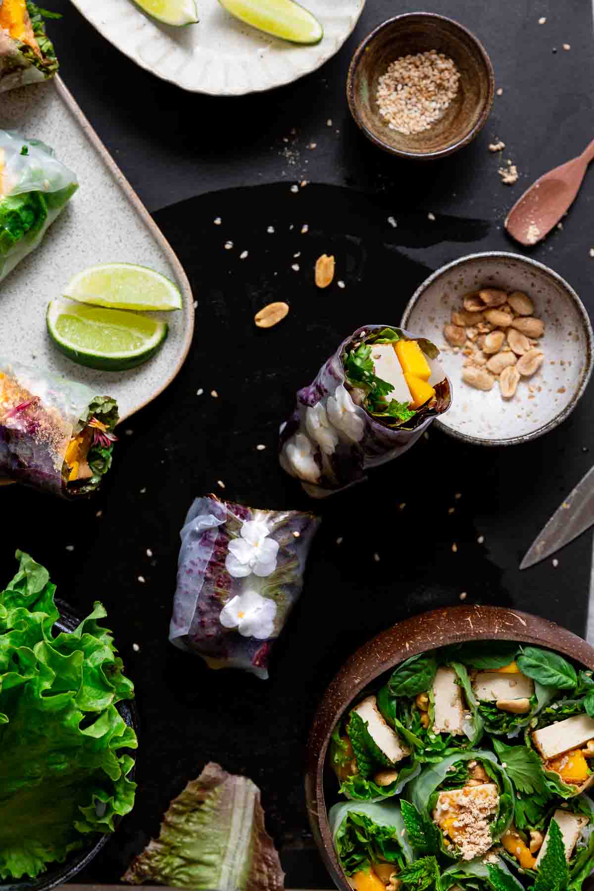 Beautiful summer rolls halved in a coconut bowl and one lined with edible flowers, halved, and leaning on a peanut bowl.