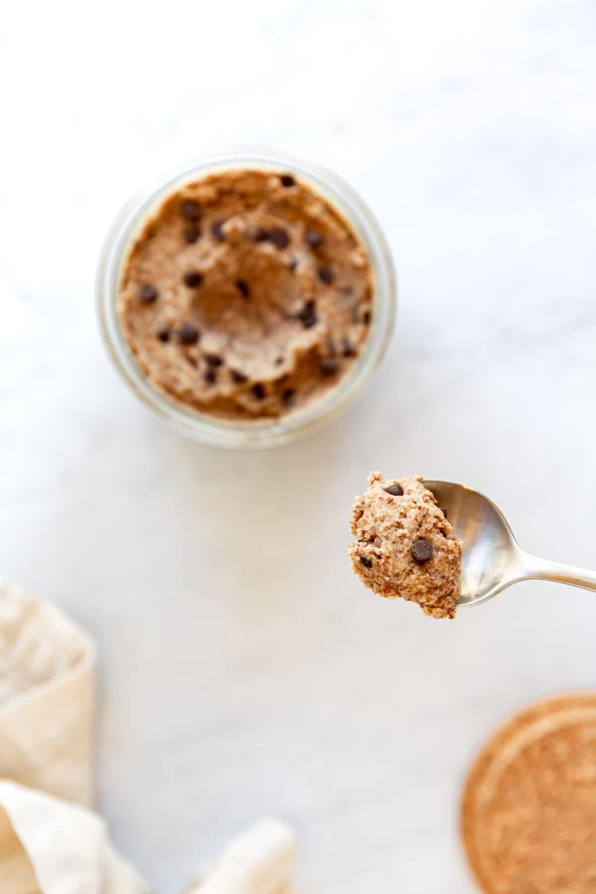 A jar of vegan gluten-free chocolate chip cookie dough on a marble surface and a spoon hovering with a spoonful of cookie dough.