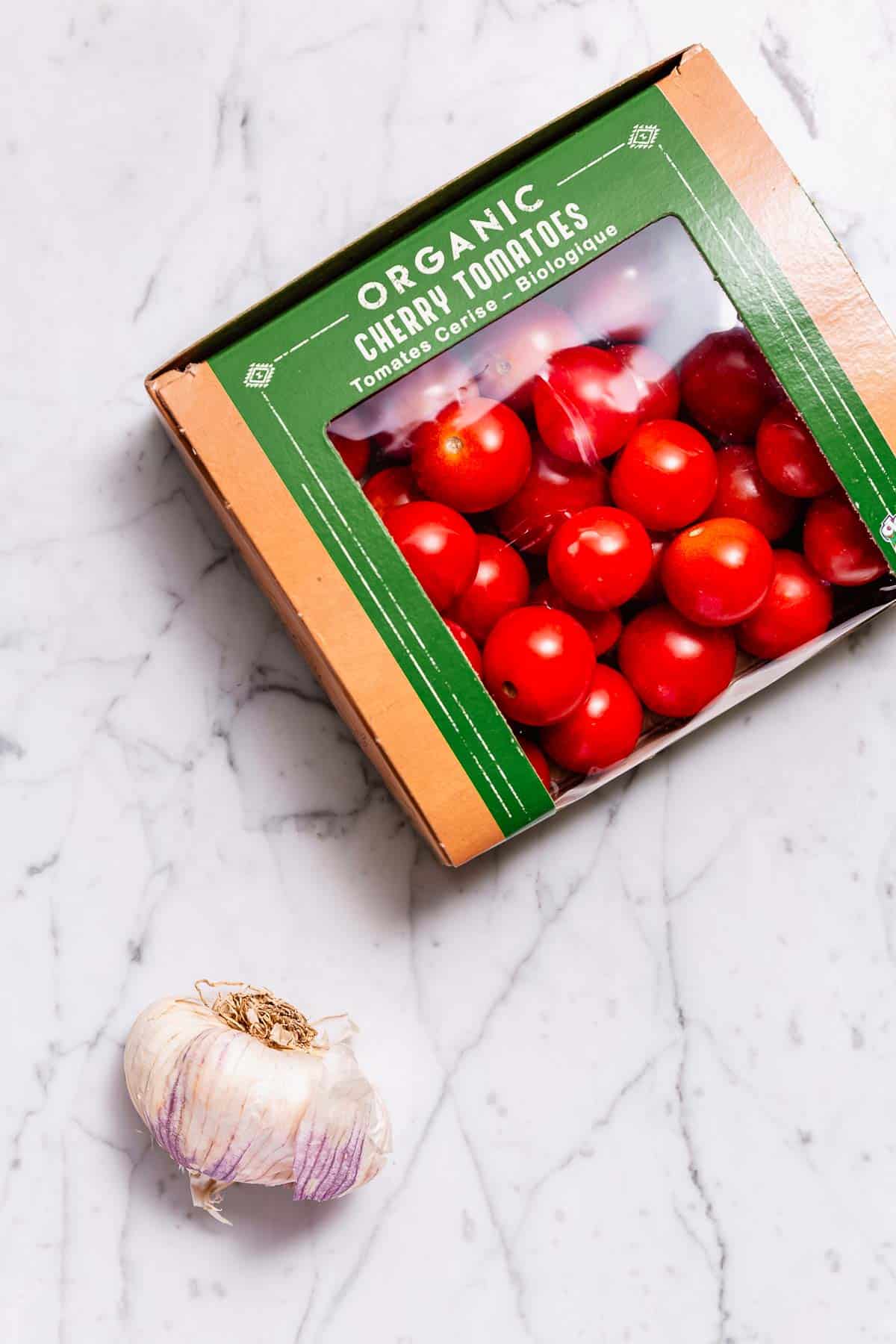 A pint box of cherry tomatoes and a head of garlic on a marble surface.