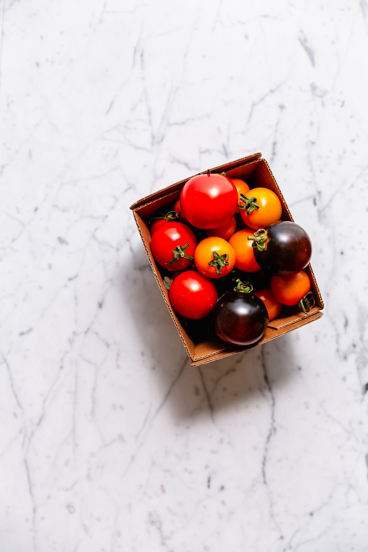 Mixed cherry tomatoes in a cardboard container on a marble surface.