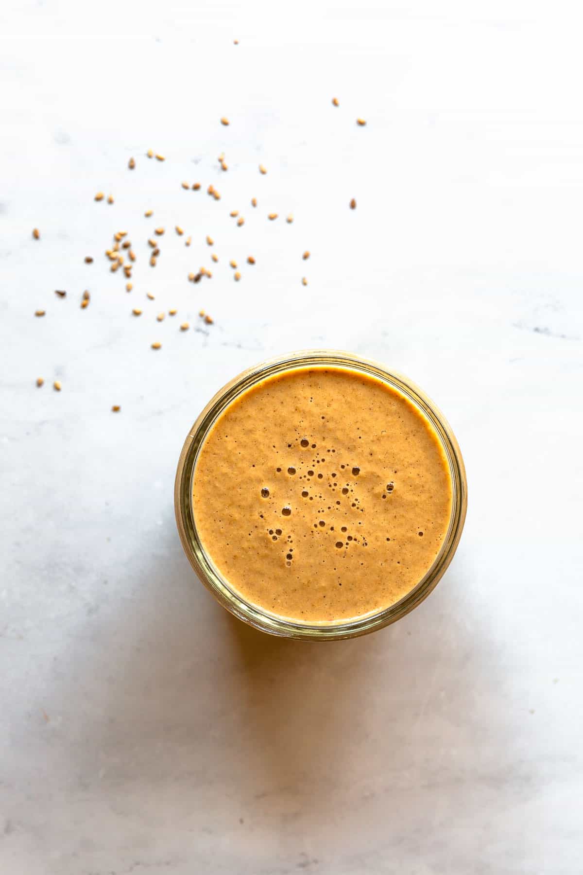 A jar of roasted sesame dressing on a marble background surrounded by scattered sesame seeds.