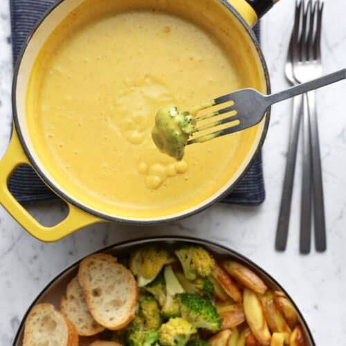 Vegan cheese fondue in a yellow pot, with a dipped piece of broccoli hovering over it.