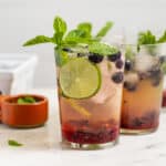 Glasses of blueberry mojito mocktails with a small bowl of fresh mint and a lime wedge.